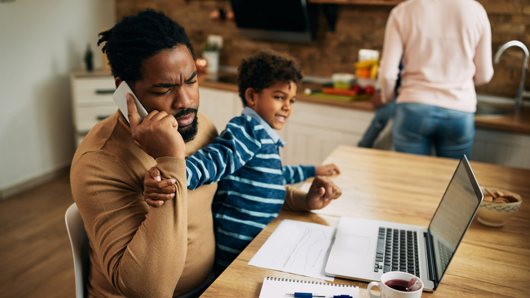 African American working father talking on the phone while his son is trying to get his attention and play with him at home.